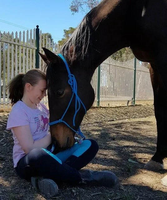 A young person sitting quietly on the grass beside a resting horse